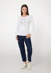 Balta ilgomis rankovėmis palaidinė su deimantais akcentuotu "GUESS" logotipu, derinama su tamsiai mėlynomis džinsomis ir baltomis sportbačiais. Modelis stovi neutralioje fone.