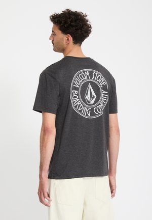 Homme aux cheveux bouclés portant un t-shirt gris foncé avec le logo "Volcom Stone Boarding Company" dans le dos, assorti à un short de couleur claire.