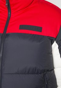 Padded bunda s červenou horní částí, námořnickým tělem a bílými akcenty. Černá logo nášivka na hrudi. Zapínání na zip a elastické manžety.