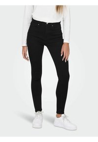 Femme portant un jean skinny noir, un haut à manches longues blanc et des baskets blanches, debout devant un fond uni.