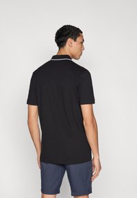 Polo noir avec un col blanc contrastant, manches courtes et texture lisse, porté avec un short bleu marine. La vue montre le dos.