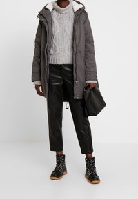 Parka grigia con cappuccio foderato in pelliccia, maglione grigio lavorato a maglia, pantaloni neri in ecopelle e stivali neri con lacci e suole in gomma; tiene in mano una borsa nera.