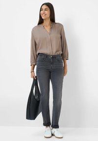 Langærmet, taupe bluse med v-udskæring og samling, parret med mørkegrå slim-fit jeans og hvide sneakers. Sort tote taske i hånden.