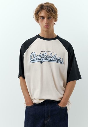 Junger Mann mit lockigem Haar, der ein schwarz-weißes "Outfielders" Baseballshirt und dunkle Jeans trägt, mit den Händen in den Taschen vor einem weißen Hintergrund steht.