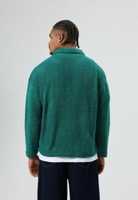 Groene fleecejas met een gestructureerd oppervlak, een omgeslagen kraag, lange mouwen en een losse pasvorm, gedragen over een wit shirt en donkere jeans.