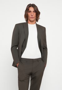 Brown tailored suit jacket with a double-breasted design and black buttons, paired with a white crew-neck t-shirt and matching trousers.