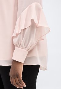 Αριστερό χέρι ατόμου που φορά διάφανο ανοιχτό ροζ πουκάμισο με πλισέ μανίκι και μανσέτα, σε συνδυασμό με μαύρο παντελόνι, πάνω σε λευκό φόντο.