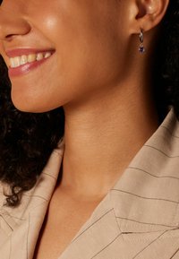 Zilveren hoepel oorbel met een ronde paarse edelsteen die hangt. Close-up van het oor, met een nadruk op de gladde huid en het beige collared kledingstuk met streepjes.