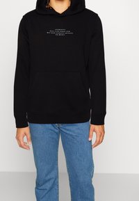 Felpa nera con tasca a canguro, con testo bianco sul petto. Tessuto morbido, polsini a coste e cappuccio con cordino. Indossata con jeans blu.
