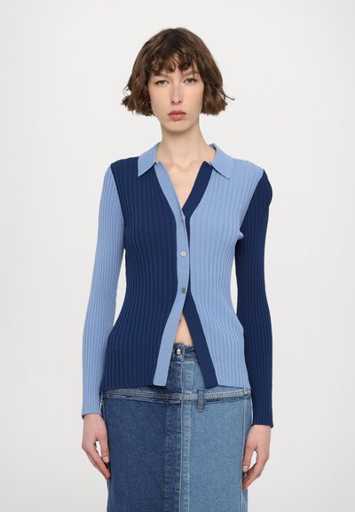 Cardigan cu model cu nervuri, caracterizat prin culori albastre în două nuanțe, cu un decolteu butonat și guler. Formă ajustată, cu mâneci lungi și o deschidere la tiv.