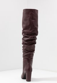 Bota de cuero burdeos sobre la rodilla con detalles arrugados, tacón alto y textura suave, mostrando el cierre de cremallera en la parte posterior.