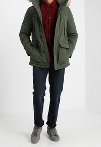 Parka matelassée verte avec capuche doublée de fausse fourrure, deux poches à rabat à l'avant, boutons, portée par-dessus une chemise bordeaux à boutons et un jean foncé. Baskets grises.