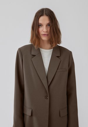 Giovane donna con capelli castani mossi fino alle spalle indossa un grande blazer marrone sopra una maglietta bianca, in piedi di fronte a uno sfondo grigio chiaro uniforme.