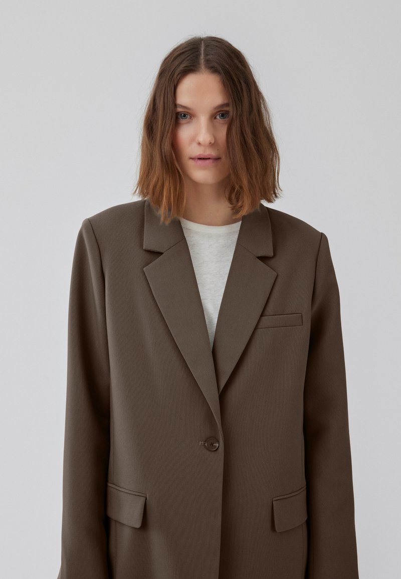 Jonge vrouw met schouderlang golvend bruin haar draagt een oversized bruine blazer over een witte top, staande tegen een effen lichtgrijze achtergrond.