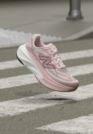 Zapatilla de correr New Balance rosa claro en el aire sobre un paso de cebra en una calle asfaltada.
