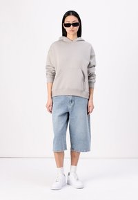 Sweat à capuche gris clair avec une poche frontale, associé à un short en jean bleu clair jusqu'aux genoux et des baskets blanches. Le modèle porte des lunettes de soleil noires.