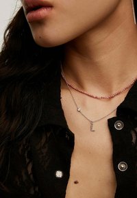 Collares de plata con capas, con un colgante en forma de letra "L" y un choker de cuentas rosas. Camisa de encaje negro con detalles de botones plateados.