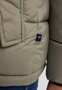 Giacca puffer beige con design a trapunta cucita e un piccolo logo nero nella parte inferiore sinistra, abbinata a jeans di denim.