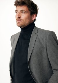 Blazer a quadri grigi sopra un dolcevita nero. Il blazer presenta un colletto a punta e una chiusura con un solo bottone, realizzato in un tessuto testurizzato.