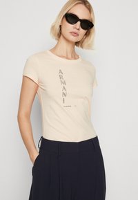 Φανελάκι σε ανοιχτό ροδάκινο, εφαρμοστό, με κοντά μανίκια και χαρακτηριστικό γραφικό λογότυπο από σκούρες ριγέ στρας. Συνδυάζεται με πλισέ σκούρο ναυτικό παντελόνι.