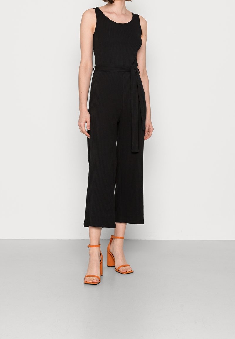 Mono negro de canalé con pantalones de pierna ancha, cintura ajustada y escote redondeado, combinado con sandalias de talón bloque con tiras en color naranja.