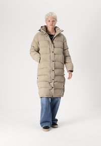 Beige pūkains mētelis ar kapuci, ar quiltotu dizainu, spiedpogām un sānu kabatām, savienots ar platām ziliem džinsiem un melnām kurpēm.