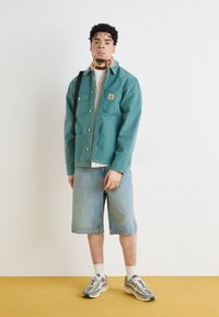 Veste en toile teal avec une poche poitrine, associée à un short en denim bleu clair. Le mannequin porte des baskets à motifs et des chaussettes blanches sur un fond jaune.