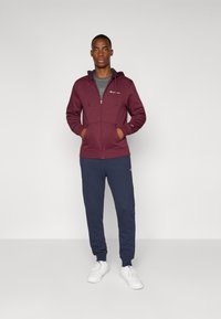 Bordeaux zip-up hoodie met zakken aan de voorkant en logo, gecombineerd met marineblauwe joggingsbroek. Beide gemaakt van zachte stof; witte sneakers maken de outfit af.