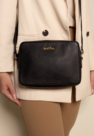 Black leather crossbody bag with a textured finish, featuring a gold-toned logo and a strap, held next to beige clothing.