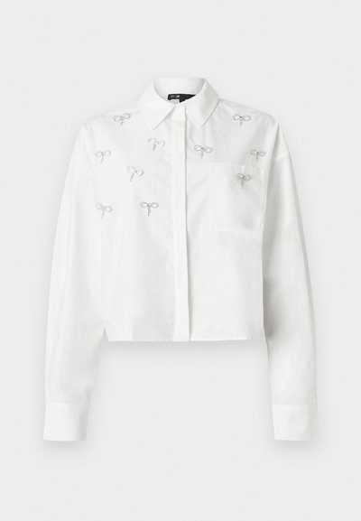 Λευκό cropped πουκάμισο με μακριά μανίκια, γιακά, μπροστινή τσέπη, κούμπωμα με κουμπιά και μικρά διακοσμητικά σε σχήμα ασημένιου φιόγκου διάσπαρτα μπροστά.