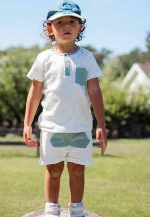 Hvid kortærmet skjorte med en teal lomme og knaplukning, parret med hvide shorts med en teal akcentlomme. Barnet bærer en matchende kasket.
