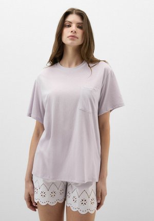 Kvinde iført en løs lys lavendel T-shirt med brystlomme og hvide shorts med hulmønster mod en ensfarvet hvid baggrund.