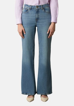 Vrouw draagt lichtblauwe hoge taille flare jeans, roze geruit overhemd met lange mouwen en beige schoenen met spits toelopende neus, staand met handen langs het lichaam.