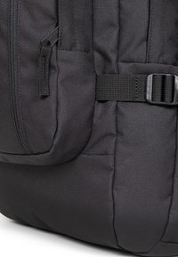 Black backpack with a textured fabric, featuring a front zippered pocket and a side strap buckle for secure closure.