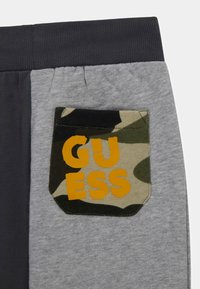 Pantalons de survêtement gris avec une taille sombre, dotés d'une poche camouflage avec "GUESS" imprimé en orange. Design texturé et décontracté.