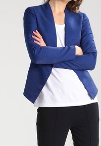 Blazer bleu à texture lisse, ouvert sur le devant et sans boutons. Associé à une chemise blanche en dessous et un pantalon noir.