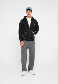 Svart zip-up hettegenser med grå sømdetaljer, kombinert med grå jeans og hvite sneakers. Modellen har på seg en lyseblå caps.