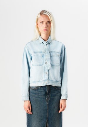 Giacca di denim blu chiaro con due tasche frontali, chiusura a bottoni e un design leggermente accorciato. Abbinata a una gonna di denim blu scuro.