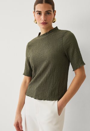 Kvinde iført tekstureret olivengrøn kortærmet top og hvide bukser, med guldringe i ørerne, stående mod en hvid baggrund.