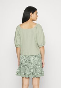 Top de linho verde com mangas bufantes, combinado com uma saia com folhos com um padrão de folhas pretas e verdes. Vista traseira mostrada.