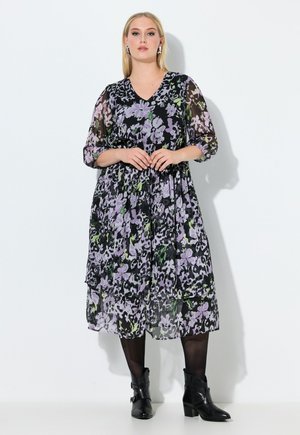 Frau trägt ein schwarzes Midikleid mit Blumenmuster und transparenten Ärmeln, schwarze Strumpfhosen, schwarze Stiefeletten und silberne Ohrringe vor weißem Hintergrund.