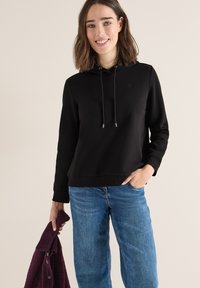Sudadera negra con cordones, puños y dobladillo acanalados, combinada con jeans de color azul claro. El modelo sostiene un cárdigan burdeos.