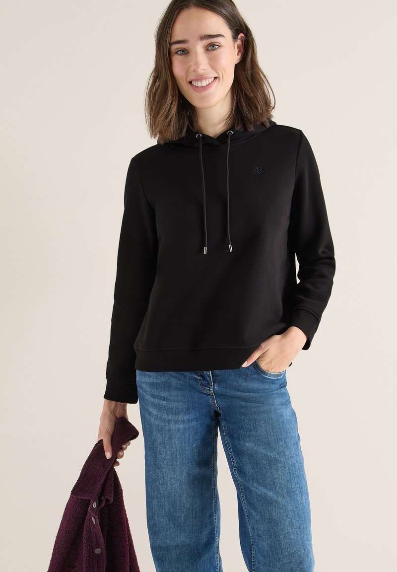 Sudadera negra con cordones, puños y dobladillo acanalados, combinada con jeans de color azul claro. El modelo sostiene un cárdigan burdeos.