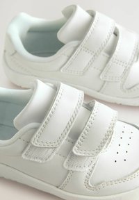 Baskets blanches en matériau synthétique lisse avec deux brides Velcro, bout perforé et semelle en caoutchouc texturée.