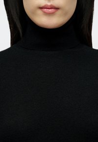 Close-up do rosto inferior e pescoço de uma pessoa a usar um suéter de gola alta preto, com os lábios fechados e cabelo liso e escuro.