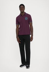 Άνδρας που στέκεται ντυμένος με ένα μωβ πόλο μπλουζάκι με μπλε λεπτομέρειες και μαύρο παντελόνι, ποζάροντας σε έναν απλό ανοιχτό φόντο.