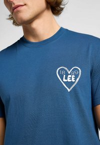 Blå bomullströja med rund halsringning. Har en vit hjärt grafik med texten "1889 LEE". Mjuk känsla, korta ärmar.