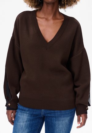 Pull en tricot marron avec un col en V et une coupe ample. Dispose de manches larges avec des accents noirs et des détails de boutons aux poignets.