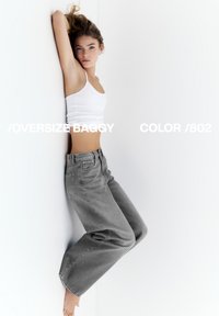 Ung kvinde i hvidt tanktop og oversize grå jeans læner sig op ad en hvid væg, ene arm løftet, barfodet, med teksten "OVERSIZE BAGGY COLOR /802" over billedet.