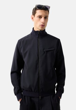 Ung mand iført sort lynlåstrøje med brystlomme og solbriller hvilende på hovedet, hænderne i lommerne, foran en ensfarvet baggrund.